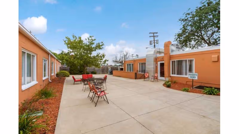 Sunny outdoor courtyard of a single-story nursing facility with patio tables and chairs between peach-colored buildings.