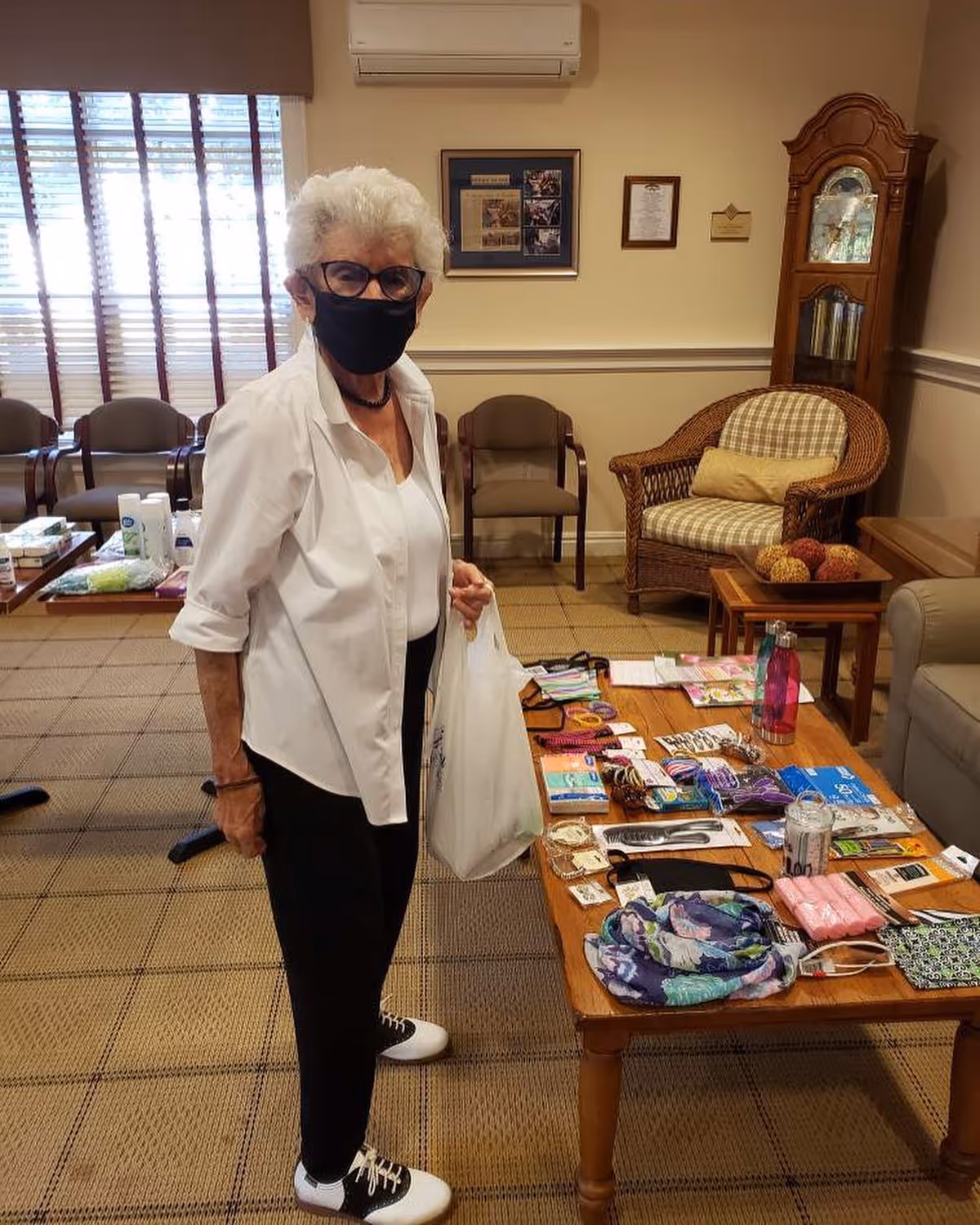 An elderly woman wearing a black face mask, glasses, white shirt, and black pants stands in a room with a table displaying various small items such as scarves, water bottles, and personal care products. The room has carpeted flooring, chairs along the wall, a wicker armchair with a cushion, a wooden grandfather clock, and framed pictures on the wall.