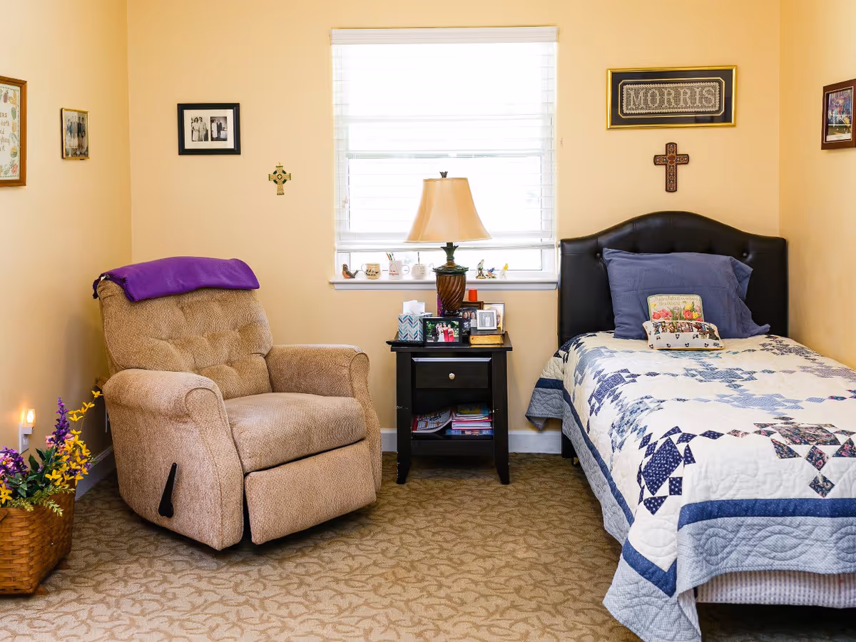 A cozy senior living bedroom featuring a single bed with a blue and white quilt, a dark cushioned headboard, and multiple pillows. Next to the bed is a black nightstand with a lamp, tissue box, framed photos, and small decorative items. To the left of the nightstand is a beige recliner chair with a purple blanket draped over the back. The room has beige walls adorned with framed pictures and crosses, a window with white blinds, and a carpeted floor with a subtle pattern. A basket with colorful artificial flowers is placed on the floor near the chair.