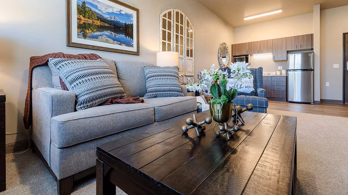 Open living room with a gray sofa and pillows, a wooden coffee table with decorative items, and a kitchenette in the background.