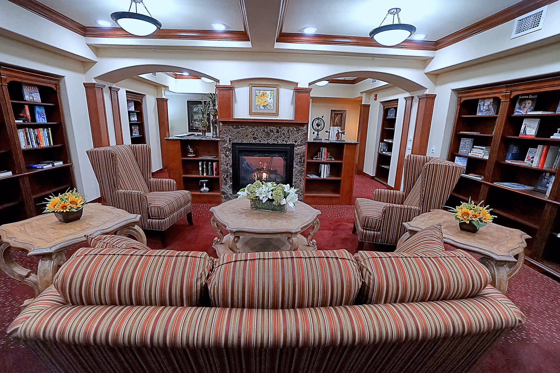 A cozy living room with a striped sofa and two matching armchairs arranged around a central coffee table with a floral centerpiece. The room features a fireplace with a marble surround, flanked by built-in wooden bookshelves filled with books and decorative items. The ceiling has recessed lighting and two hanging light fixtures, with crown molding along the edges. The carpet is red, complementing the warm wood tones of the furniture and shelves.