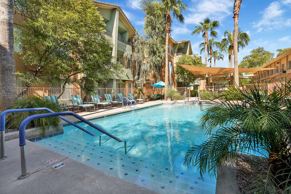 Outdoor swimming pool in a courtyard surrounded by lounge chairs, palm trees, and multi-story residential buildings.