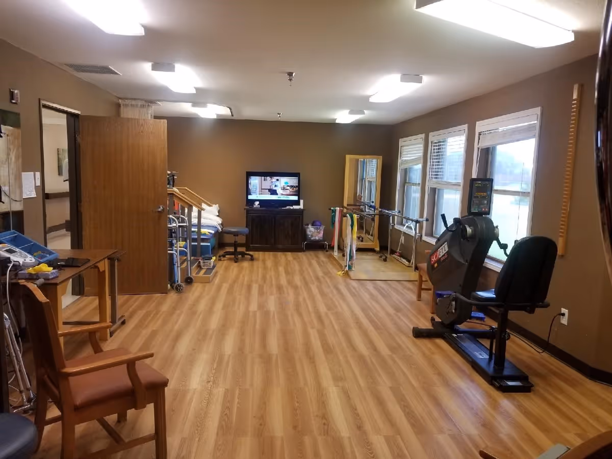 A rehabilitation room with wooden flooring and brown walls, featuring exercise equipment including a stationary recumbent bike, parallel bars for walking exercises, a TV on a cabinet, chairs, and various therapy tools. There are windows on the right side letting in natural light.