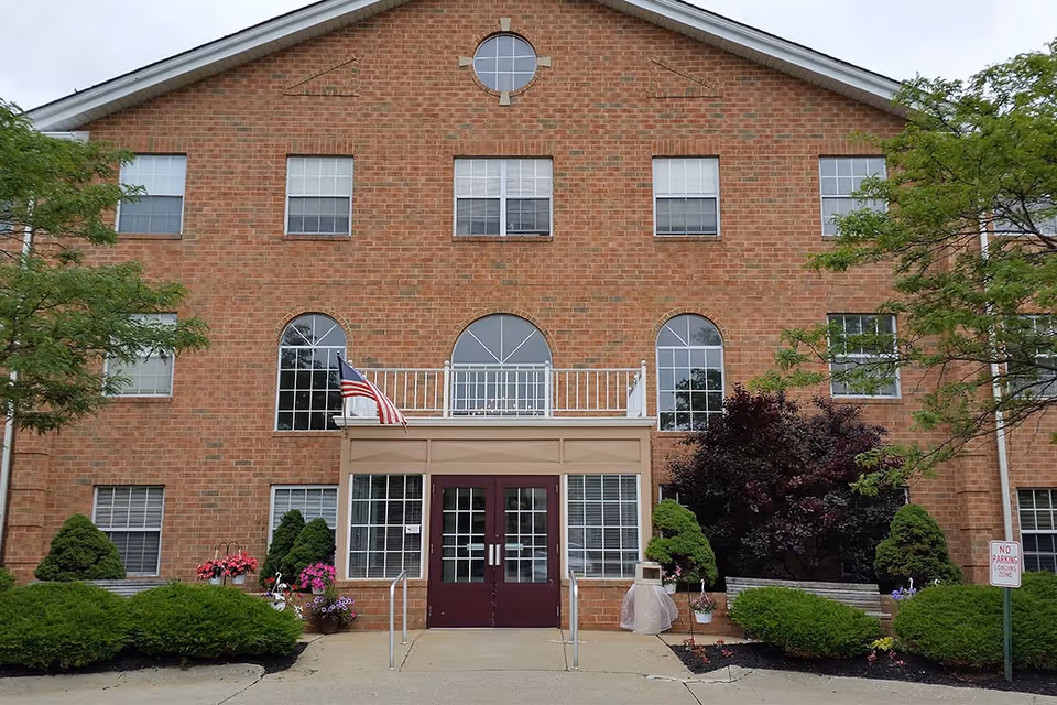 Front exterior view of a three-story brick building with multiple windows, a central entrance with double glass doors, an American flag, and landscaped bushes and trees around the entrance.