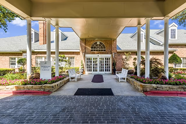 Covered main entrance of a brick senior living clubhouse with white columns, benches, flower beds, and double glass doors.