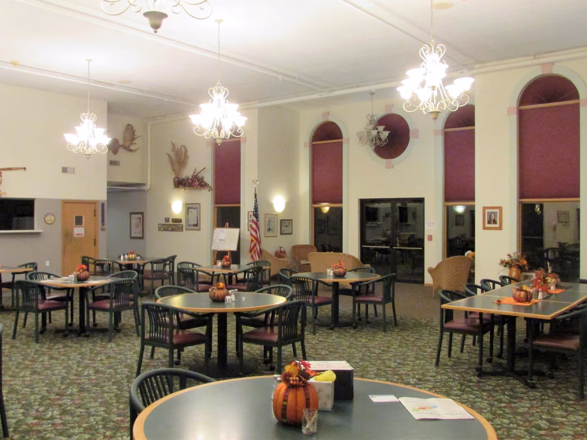 A spacious dining room in Eastland Retirement Community with multiple round and rectangular tables surrounded by chairs. The tables are decorated with small pumpkin centerpieces and autumn-themed decor. The room features high ceilings with ornate chandeliers, large arched windows with red blinds, and an American flag near the back wall. The carpet has a floral pattern, and there are some framed pictures and wall decorations visible.