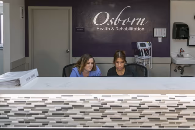 Two healthcare workers sitting behind a reception desk in a medical facility with a sign on the wall that reads 'Osborn Health & Rehabilitation'. The desk has a modern tiled front, and there is medical equipment and a sink visible in the background.