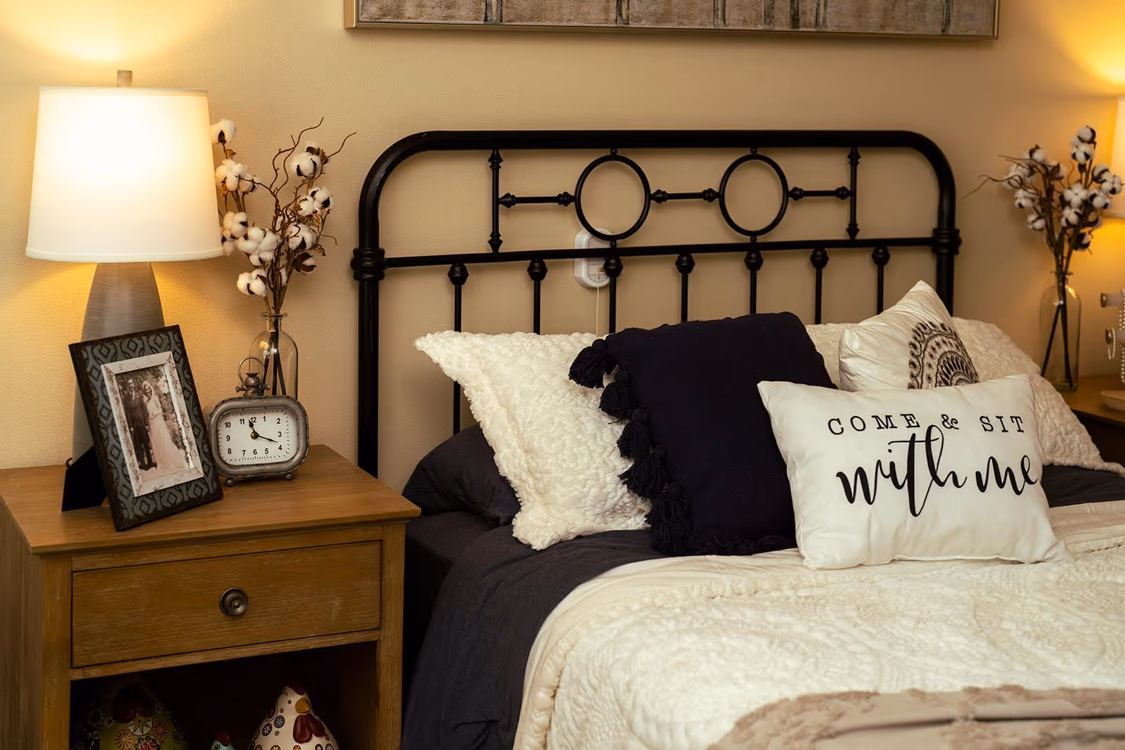 Cozy bedroom with a black metal headboard, layered pillows (one reading "Come & Sit with me"), and a wooden bedside table holding a lamp, alarm clock, and framed photo.