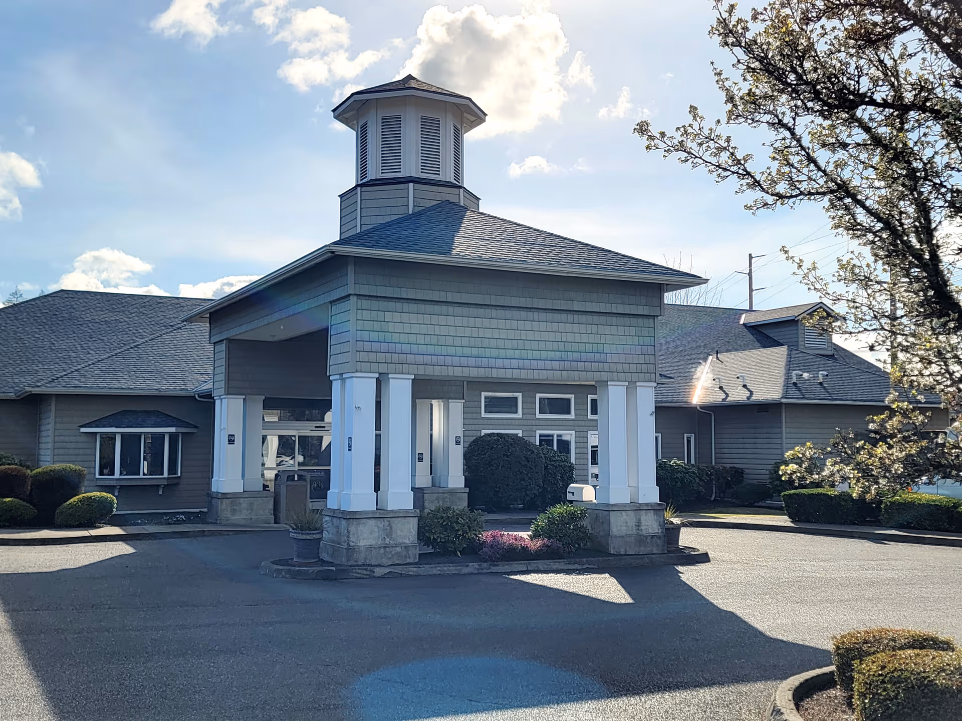 Exterior view of Shelton Health and Rehabilitation Center showing the main entrance with a covered drop-off area supported by white columns, surrounded by landscaped bushes and trees under a partly cloudy sky.