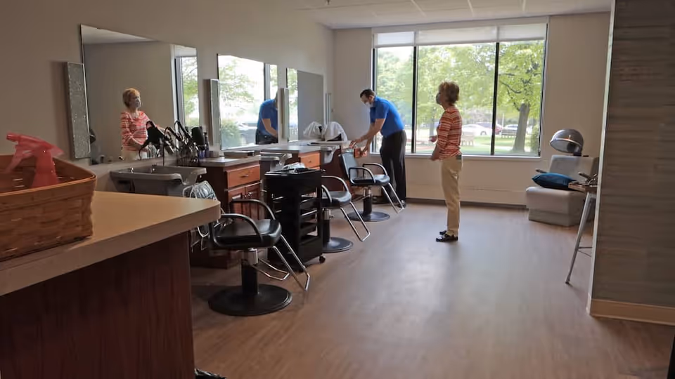 Interior salon room with styling chairs, mirrors, and a hairdresser attending to a woman near large windows.