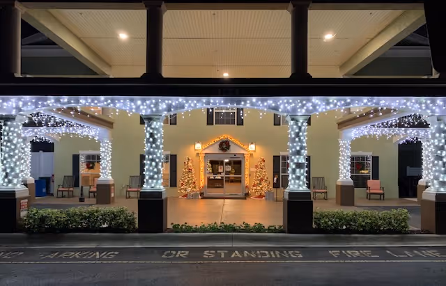 Entrance of a building decorated with white string lights wrapped around columns and hanging from the roof. Two Christmas trees with lights and ornaments flank the doorway, which has a wreath above it. Several chairs are placed along the walls under covered walkways.
