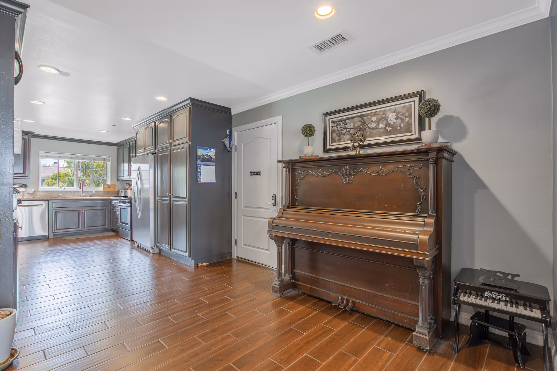 Interior view of a kitchen area with dark cabinetry, stainless steel appliances, and a window above the sink. Adjacent to the kitchen is a wooden upright piano with decorative carvings, topped with two small potted plants and a framed floral artwork on the wall above it. The floor is covered with wood-look tiles.