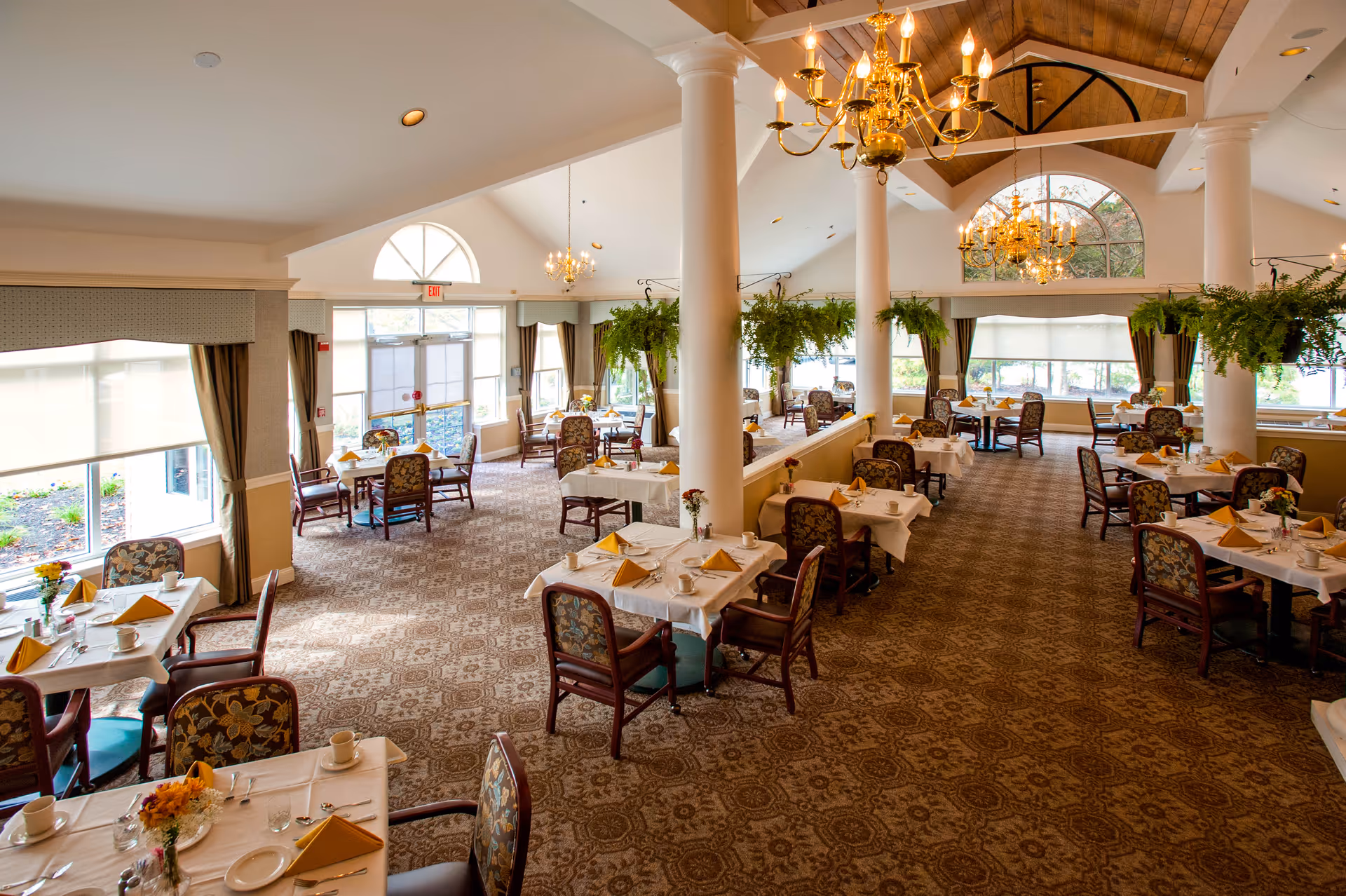 A spacious and elegant dining room in a senior living facility with multiple tables covered in white tablecloths, each set with plates, cups, utensils, and folded yellow napkins. The room features large windows with curtains, hanging plants, chandeliers, and patterned carpet flooring.