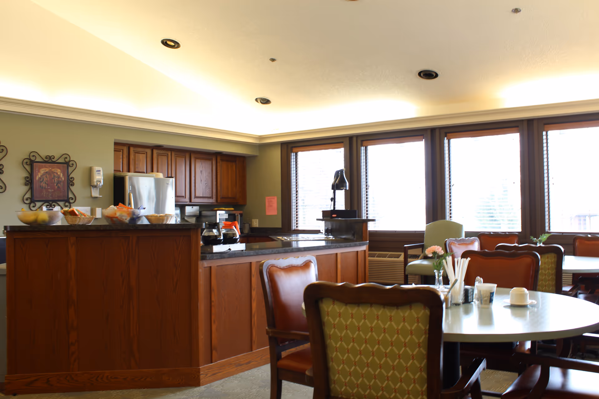 Communal dining area with round tables and upholstered chairs, a service counter with wooden cabinetry and a refrigerator, and large windows letting in daylight.