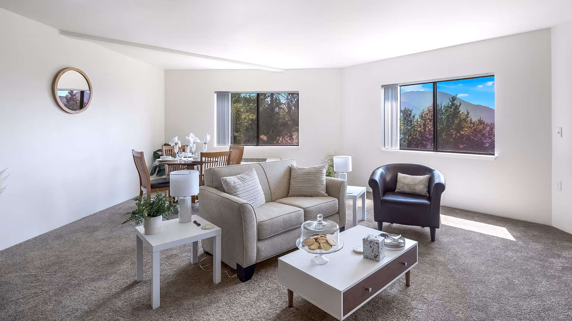 Bright living room with a sofa, armchair, coffee table and dining table beside windows showing trees and a mountain view.