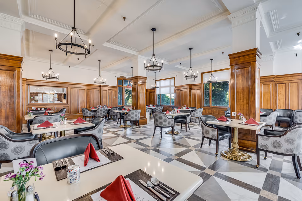 Spacious, elegant dining room with multiple set tables, patterned chairs, wood paneling and chandeliers.