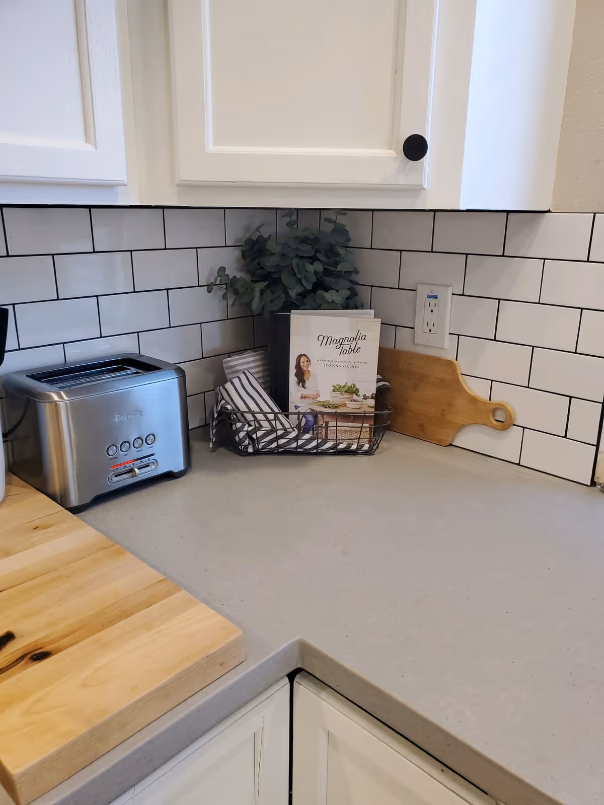 Kitchen countertop corner with a toaster, cutting boards, a cookbook in a wire basket, and white subway tile backsplash.