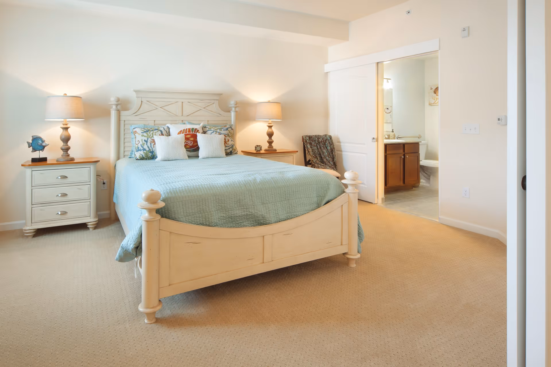 A bedroom with a light wooden bed frame and a light blue quilted bedspread. The bed has decorative pillows with sea-themed designs. There are two matching wooden nightstands on either side of the bed, each with a lamp. To the right, there is an open sliding door leading to a bathroom with a wooden vanity and a toilet visible. The room has beige carpet and light-colored walls.