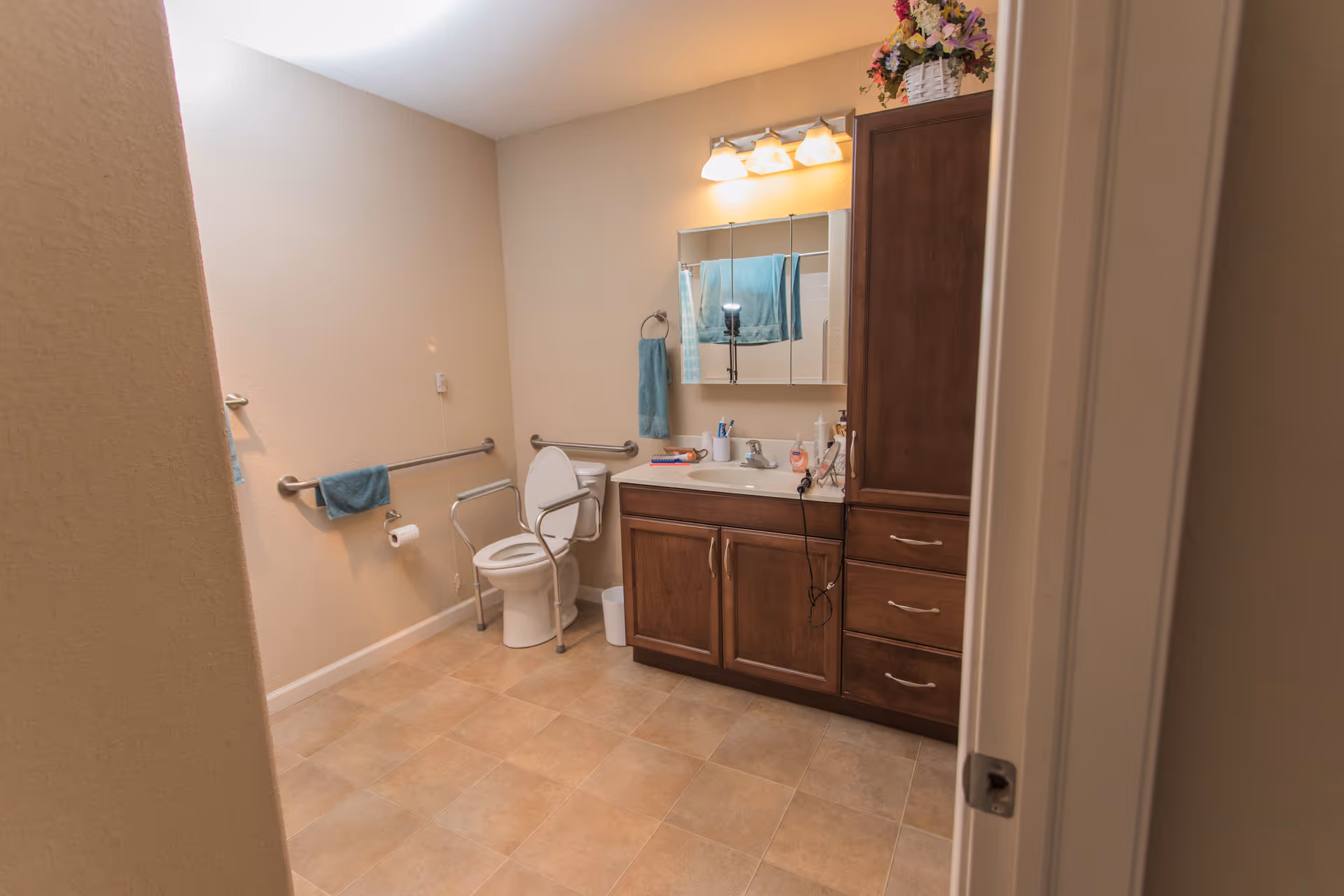Accessible bathroom with a toilet fitted with safety rails, a wooden vanity and mirror, towels, and tiled floor.