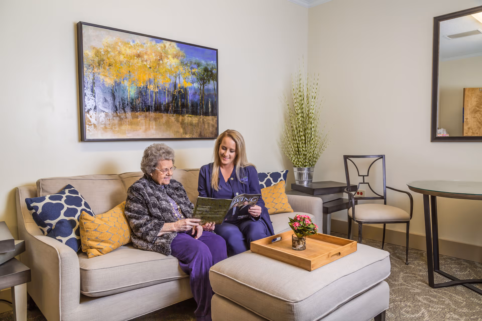 An elderly woman and a younger woman sit on a sofa in a bright lounge area reading a magazine.