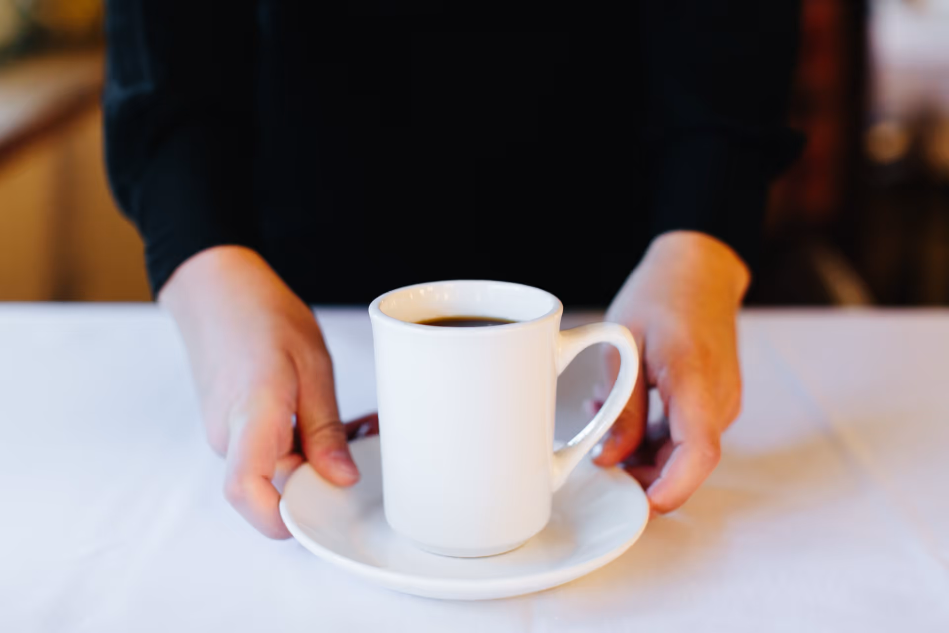 Hands placing a white coffee mug on a saucer on a white tablecloth.