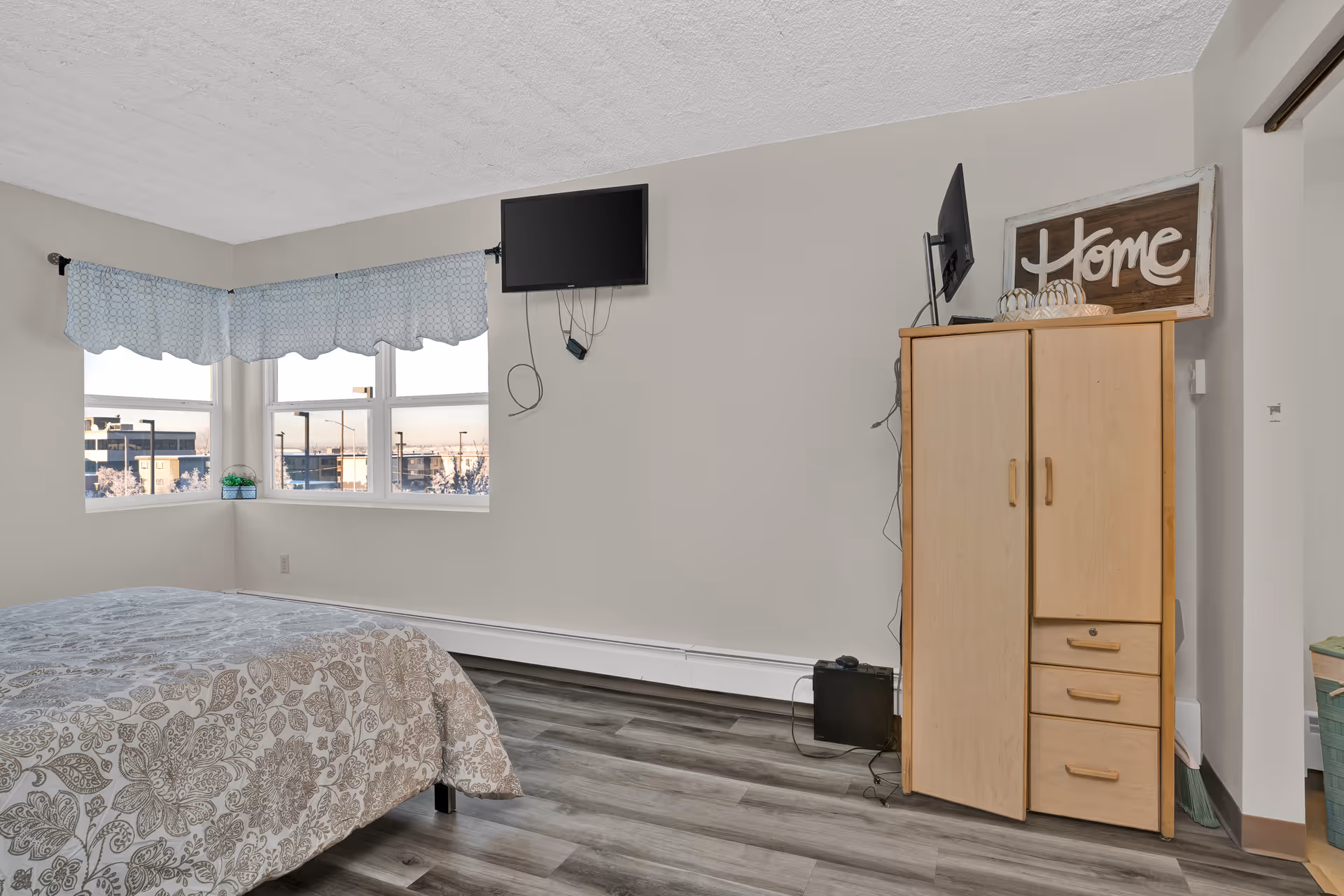 A bedroom with a bed covered in a patterned bedspread, a corner window with blue valance curtains, a wall-mounted flat screen TV, and a light wood wardrobe with a decorative 'Home' sign on top.