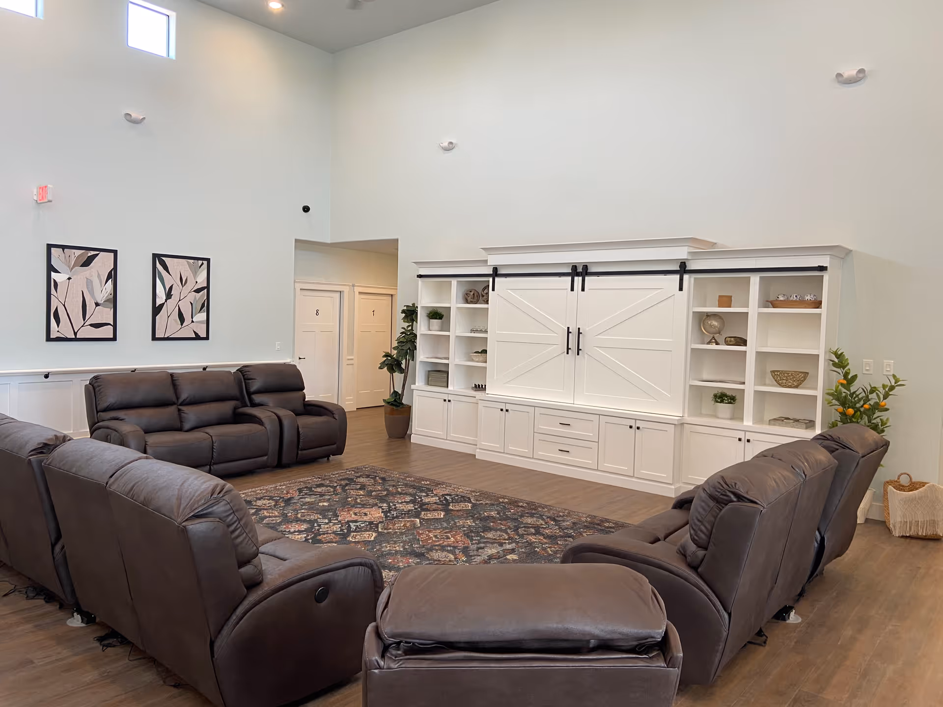 A spacious communal living room with brown reclining sofas arranged around a patterned rug facing built-in white shelving with sliding barn-style doors.