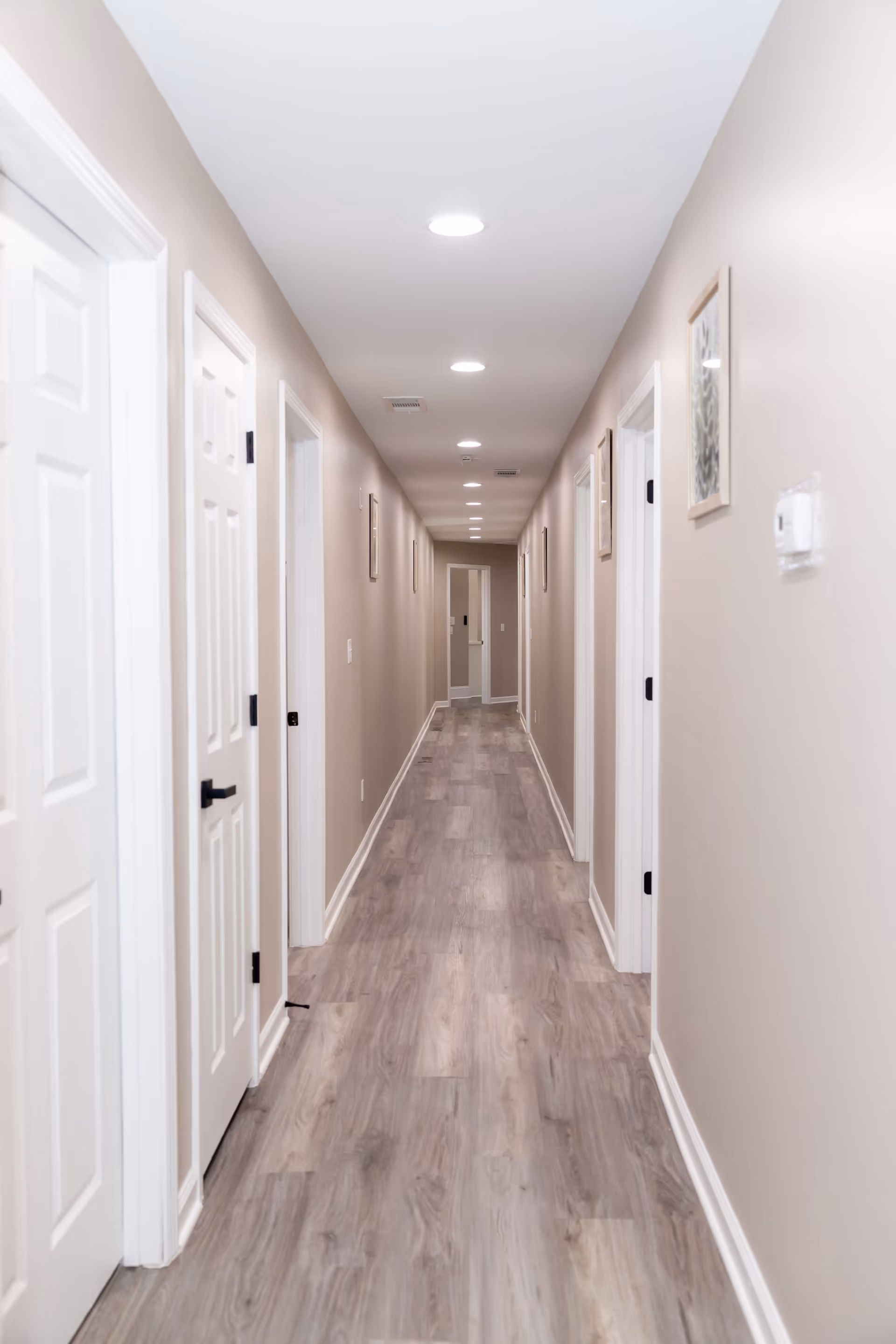 A long, narrow hallway with light beige walls and wood-patterned flooring. The hallway is lined with white doors on both sides and has recessed ceiling lights evenly spaced along the ceiling. There are framed pictures on the walls and a thermostat on the right wall.