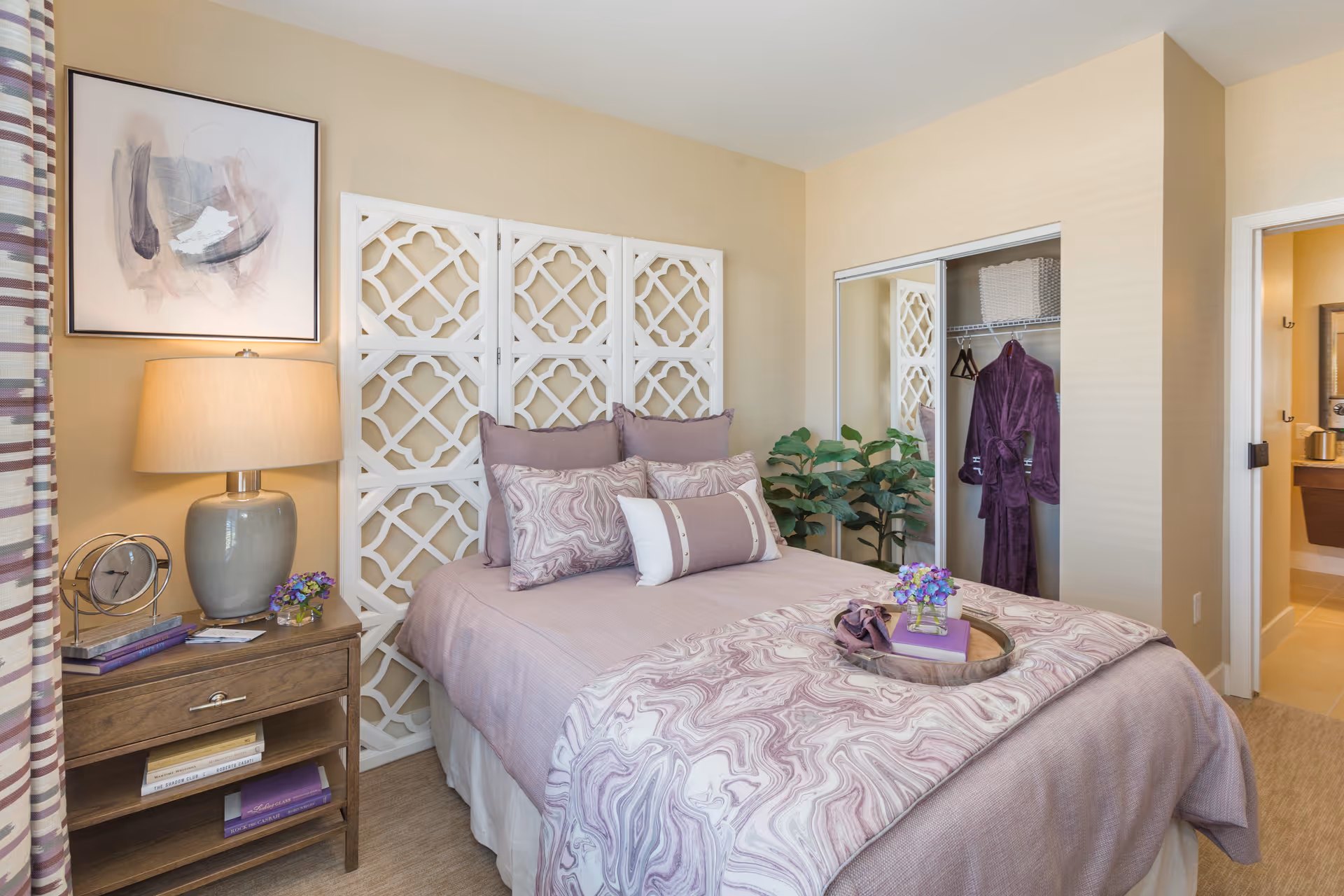 A cozy bedroom with a bed dressed in lavender and patterned bedding, decorative pillows, and a wooden tray with a book and flowers. There is a wooden nightstand with a lamp, clock, books, and a small flower vase. A white decorative room divider is behind the bed. A closet with mirrored sliding doors is visible, with a purple robe hanging inside. A plant is placed near the closet, and an open door leads to a bathroom.