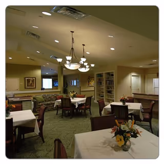 Dining room with several small tables covered in white tablecloths and floral centerpieces, chairs, a chandelier, and a seating area with shelving in the background.