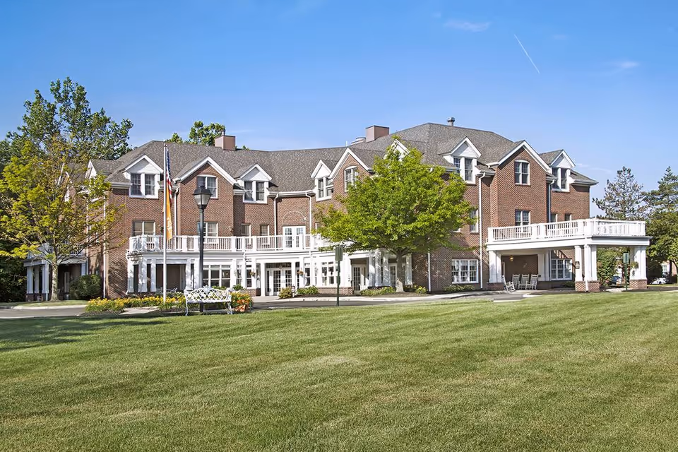 Front exterior of a large brick senior living facility with white balconies, a manicured lawn, and trees.