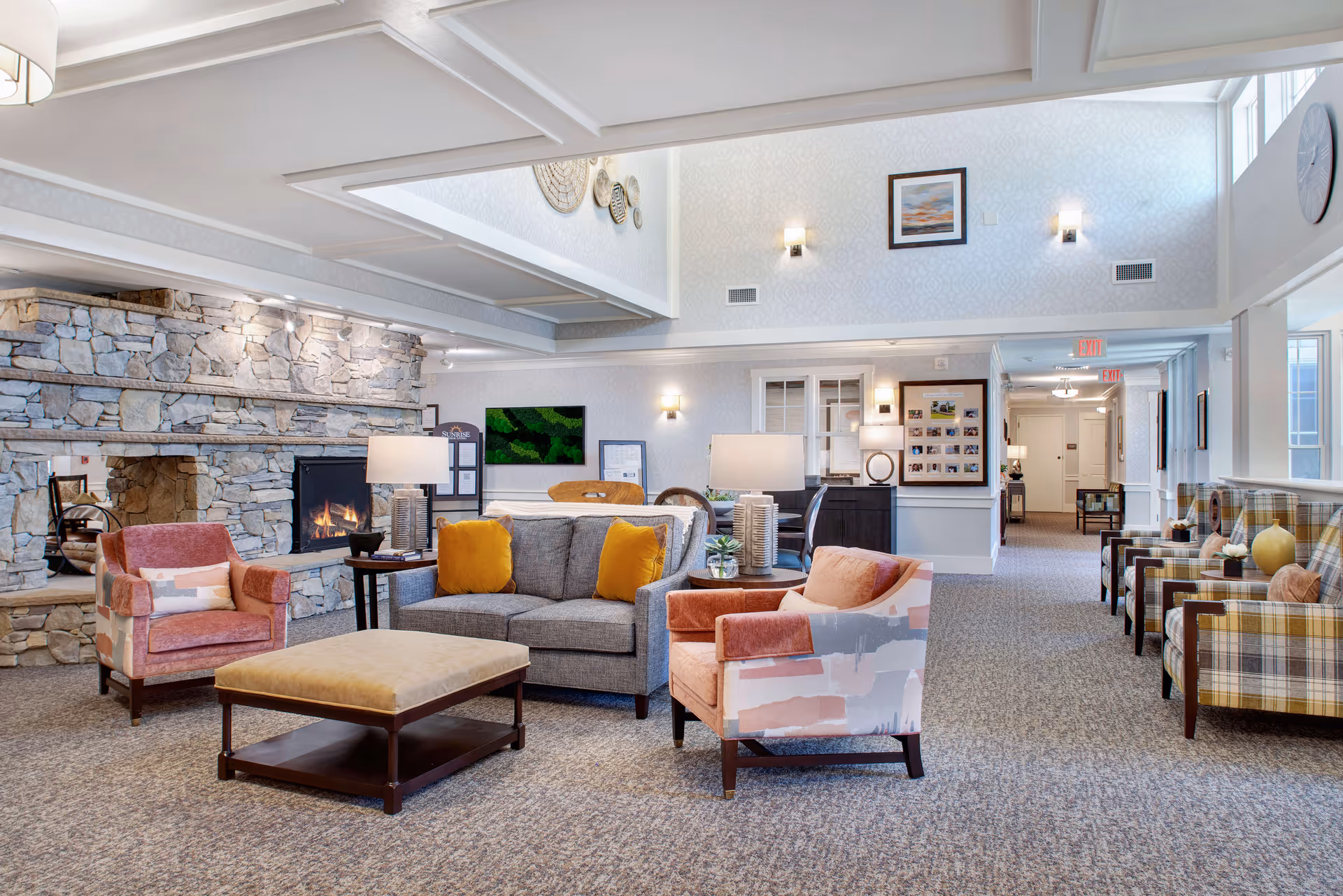 A spacious and well-lit living room area in a senior living facility featuring a stone fireplace with a fire burning, a gray sofa with yellow cushions, two patterned armchairs, a large ottoman, and several lamps. The room has high ceilings with recessed lighting, framed artwork on the walls, and a carpeted floor. There are additional seating areas with plaid chairs along the right side and a hallway leading to other rooms in the background.