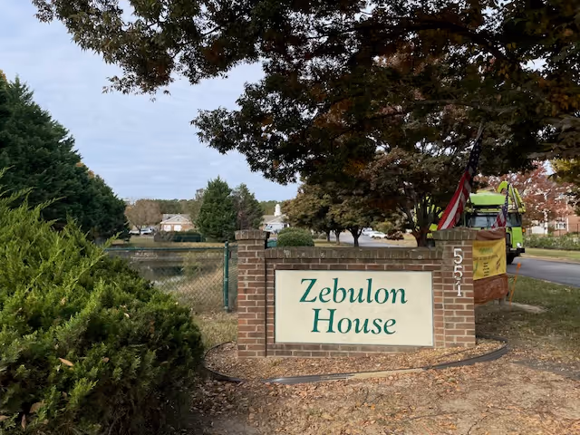 Brick entrance sign reading "Zebulon House" by a pond and tree-lined street.