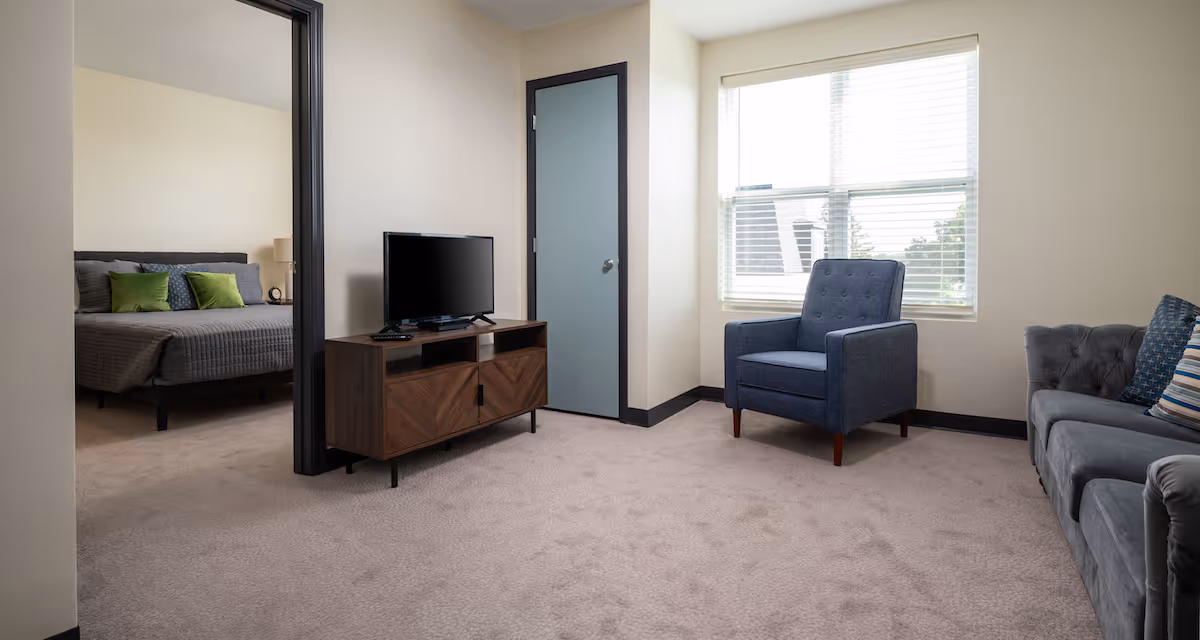 A cozy living room with a gray sofa and a blue armchair near a window with blinds. A wooden TV stand with a flat-screen television is positioned against the wall next to a closed door. Through an open doorway, a bedroom with a bed featuring gray bedding and green pillows is visible.