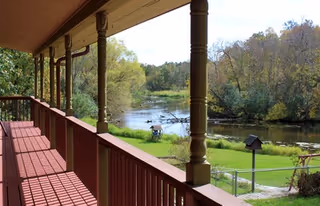 Covered wooden balcony with red railings overlooking a grassy yard and a pond surrounded by trees.