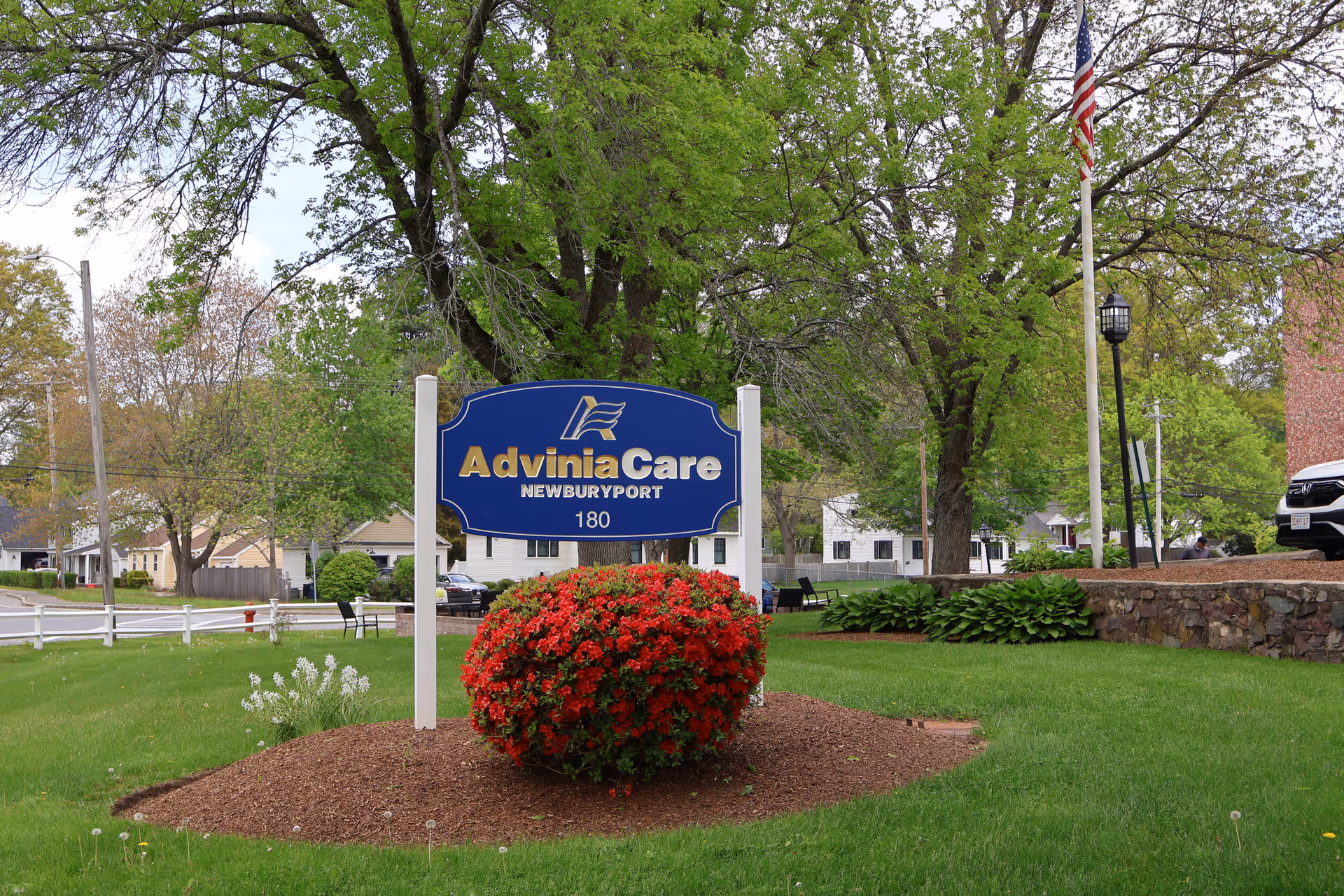 A blue and gold sign for AdviniaCare Newburyport with the number 180 below it, set in a landscaped area with green grass, a red flowering bush, and trees in the background. Nearby are a flagpole with an American flag, a stone wall, and a parked vehicle.