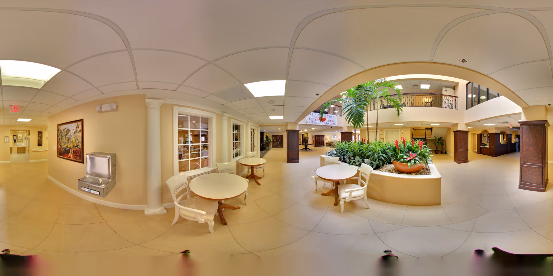 Spacious and well-lit interior common area of a senior living facility featuring a central planter with lush green plants and red flowers. There are round tables with white chairs arranged around the planter. The area has beige tiled floors, cream-colored walls, decorative columns, and a ceiling with recessed lighting. A water fountain is mounted on one wall, and a hallway extends to the left. The upper level has a balcony with railing overlooking the area below.