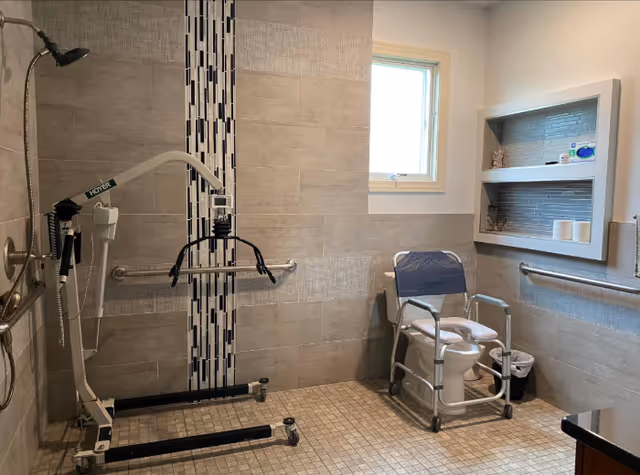 Accessible bathroom with beige tiled walls and floor, featuring a shower area with a handheld showerhead, a Hoyer lift for patient assistance, grab bars on the walls, a window, a commode chair with armrests and wheels, and a recessed shelf holding toiletries and toilet paper.