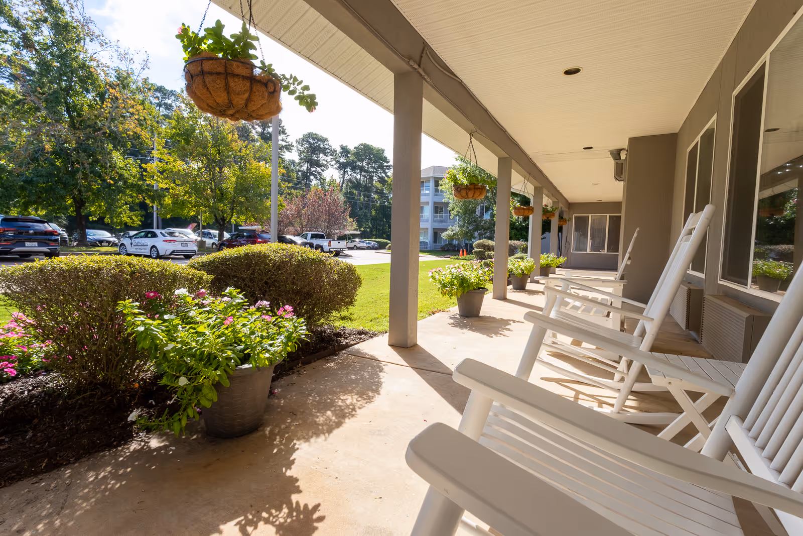 A sunny outdoor porch area at Asher Point Independent Living of Texarkana with white rocking chairs lined up along the wall. Hanging flower baskets and potted plants decorate the porch, with a view of green bushes, trees, and parked cars in the background.