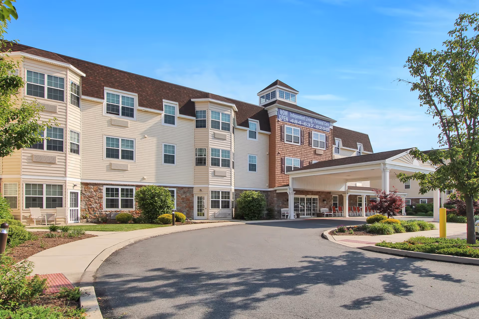 Exterior view of Keystone Villa at Fleetwood, a multi-story senior living facility with beige siding, stone accents, multiple windows, and a covered entrance driveway. The building is surrounded by landscaped greenery and trees under a clear blue sky.