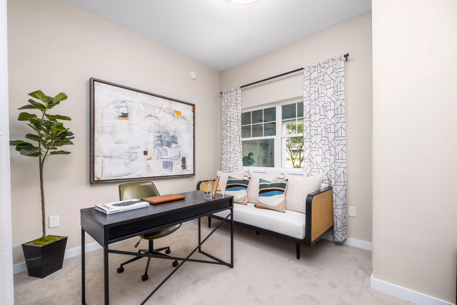 A small office or study room with a black desk and green swivel chair. On the desk are a book, a closed laptop, and a reed diffuser. Behind the desk is a modern black-framed sofa with white cushions and decorative pillows in beige and blue stripes. A large abstract painting hangs on the beige wall to the left, and a tall potted plant is in the corner. A window with patterned white curtains lets in natural light.