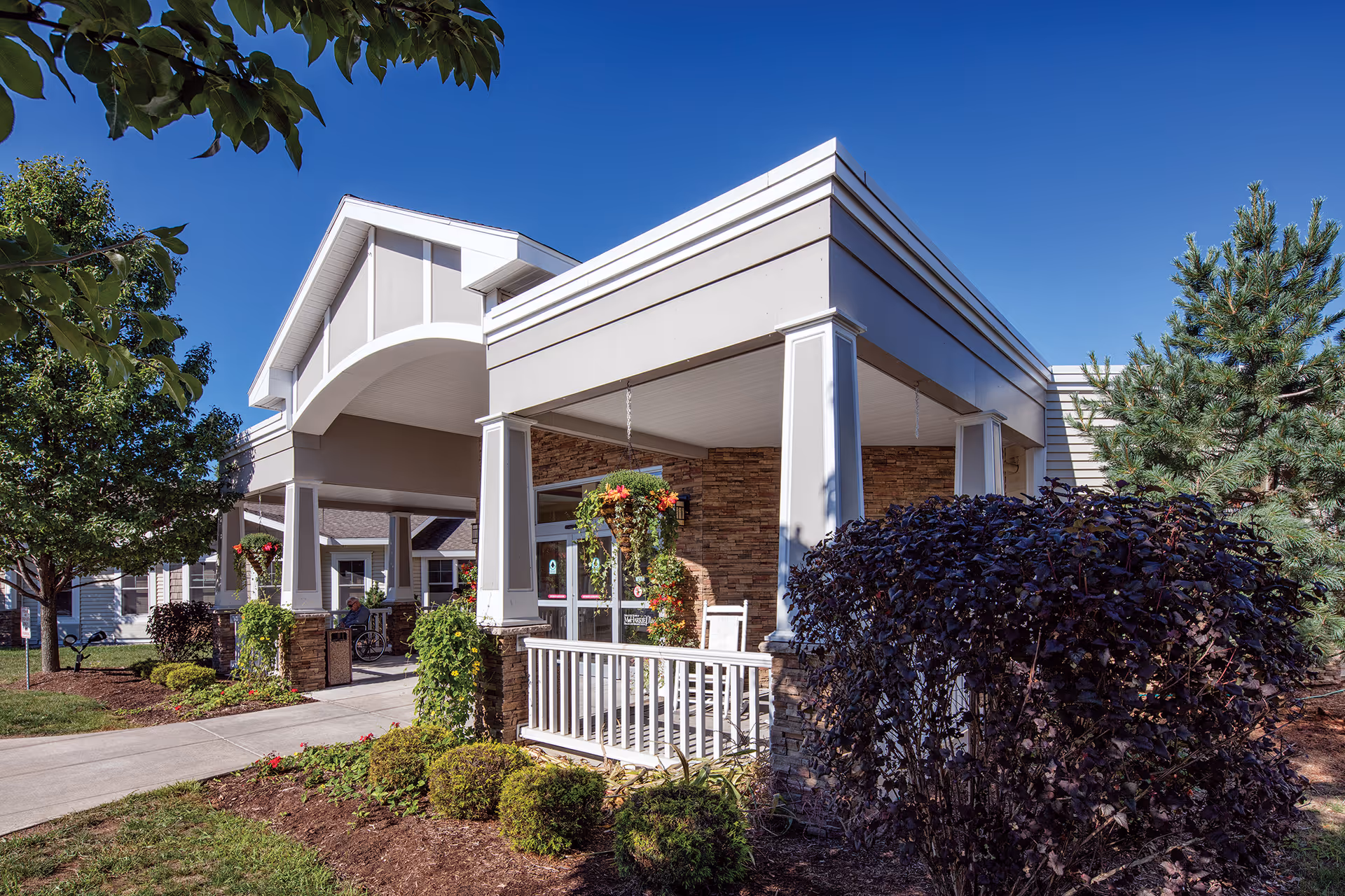 Exterior view of Mc Harrie Pointe facility entrance with a covered porch, white columns, hanging flower baskets, and well-maintained landscaping including bushes and trees under a clear blue sky.