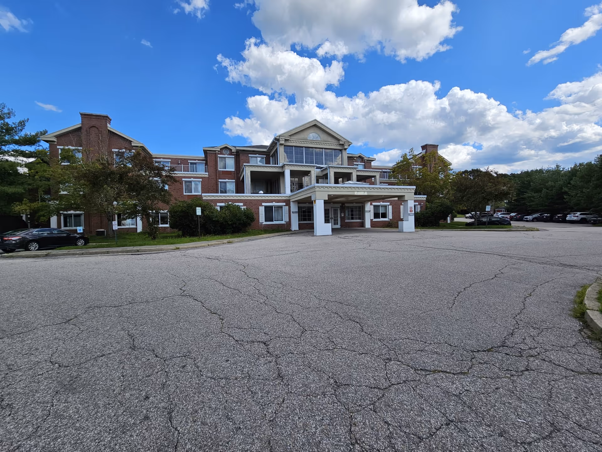 Front view of a three-story brick nursing center with a covered entrance/porte-cochère and a large cracked parking lot under a blue sky.