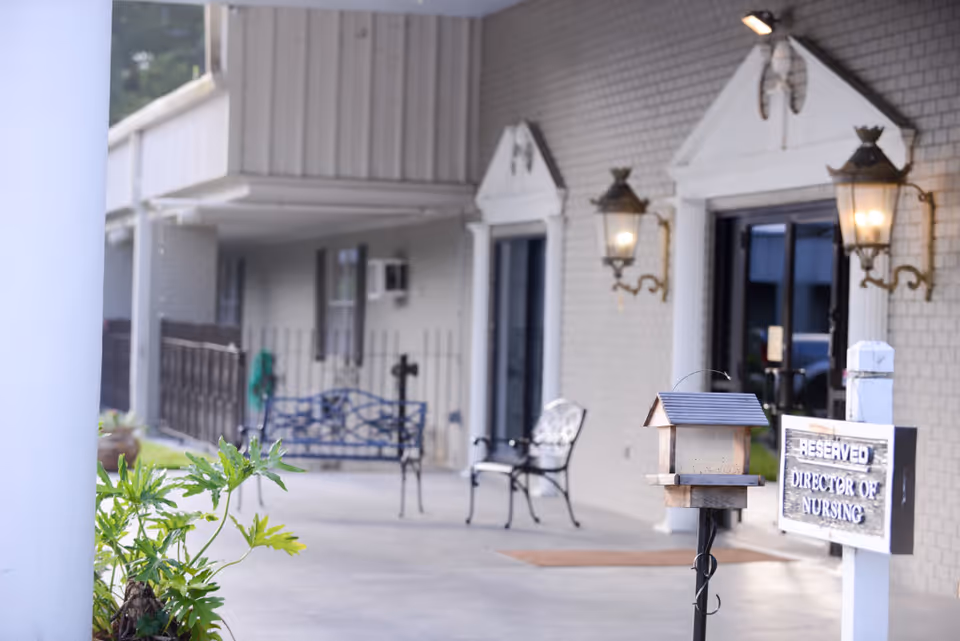 Outdoor covered walkway area at Pontchartrain Health Care Center with benches, a birdhouse on a post, and a sign that reads 'Reserved Director of Nursing'. The building exterior features brick walls, decorative wall lamps, and a glass door entrance.