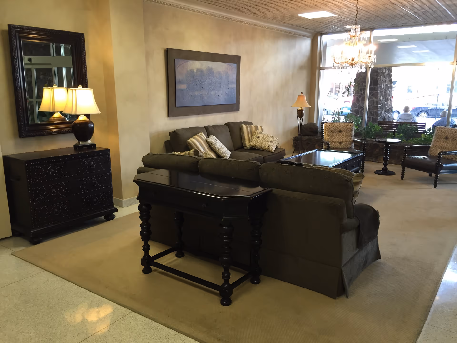 A cozy living room area with a dark green sofa, two patterned armchairs, a dark wooden coffee table, and a matching console table. There is a large mirror above a chest of drawers with a lamp on it, a framed artwork on the wall, and a chandelier hanging from the ceiling. Large windows in the background show an outdoor view with people sitting on benches.