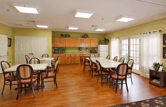 A bright dining room with multiple tables and chairs arranged neatly. The room has wooden flooring, light green walls, and a set of wooden cabinets along one wall with a refrigerator. Large windows with sheer white curtains allow natural light to fill the space. There are some plants and decorative items on top of the cabinets and a small table with a plant and framed picture near the windows.