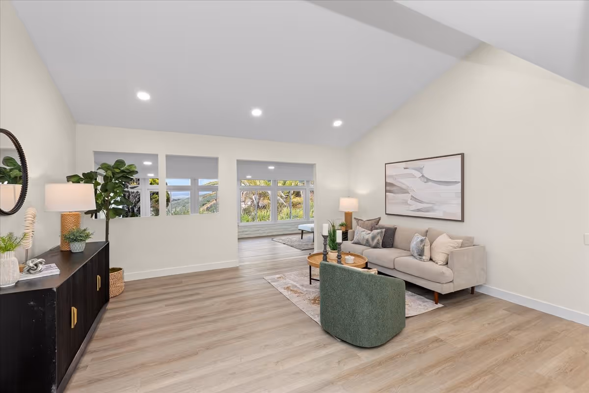A modern living room with light wood flooring, a beige sofa with multiple cushions, a green armchair, and a round wooden coffee table with decorative items. There is a black sideboard with plants and a lamp on the left side, and a large window in the background showing greenery outside. The walls are painted white, and the ceiling has recessed lighting.