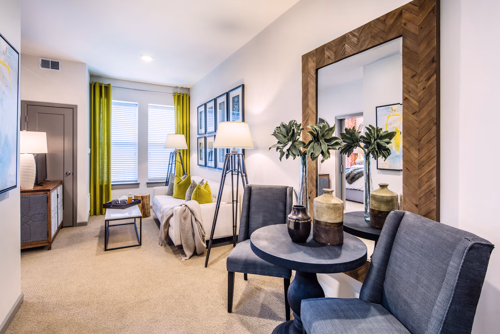 A cozy living room area with a white sofa adorned with green pillows and a beige throw blanket, two tall floor lamps, a small coffee table, and a sideboard with a lamp. There are green curtains on the windows and framed artwork on the wall. In the foreground, there is a round table with decorative vases and two gray upholstered chairs, with a large wooden-framed mirror on the wall reflecting part of a bedroom.