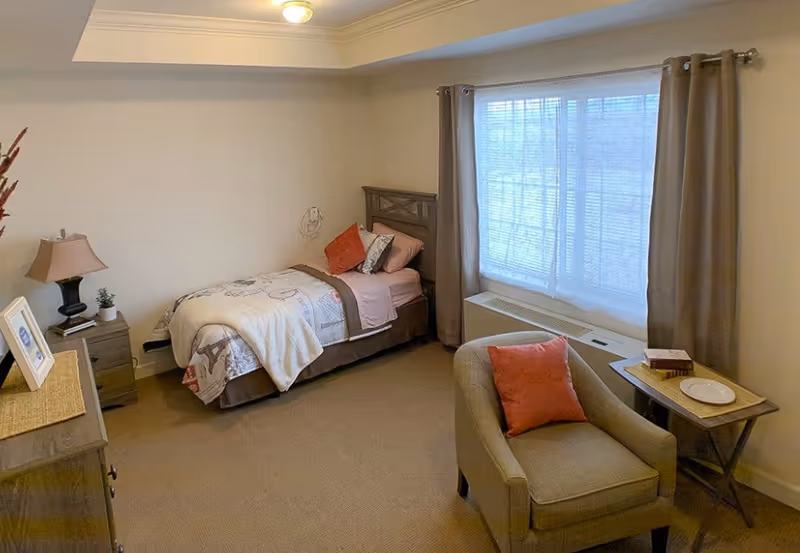 A cozy bedroom in an assisted living facility featuring a single bed with pillows and a blanket, a bedside table with a lamp and a small plant, a comfortable armchair with an orange cushion, a side table with a book and a plate, and a large window with curtains letting in natural light.