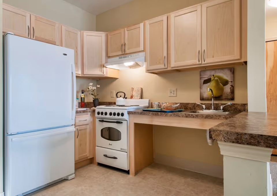 A compact kitchen with light wood cabinets, a white refrigerator, a white stove with a kettle on it, a double sink, and a countertop with a tray holding mugs and bread. There is a picture of pears on the wall above the sink.